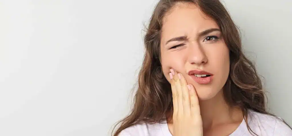A young woman holds her cheek with a pained expression, representing the need for emergency dental care due to a toothache.